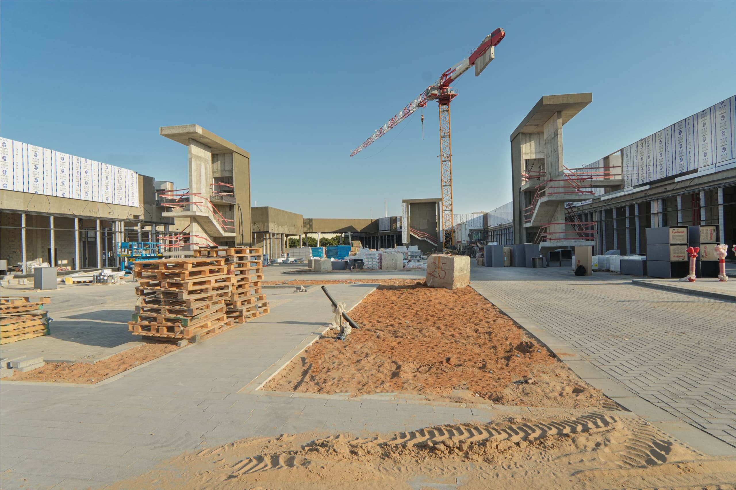 רחבת המתחם בבנייה — מגדל מדרגות בטון משני צדי הפריים, מנוף צריח במרכז ומשטחי בנייה בחזית